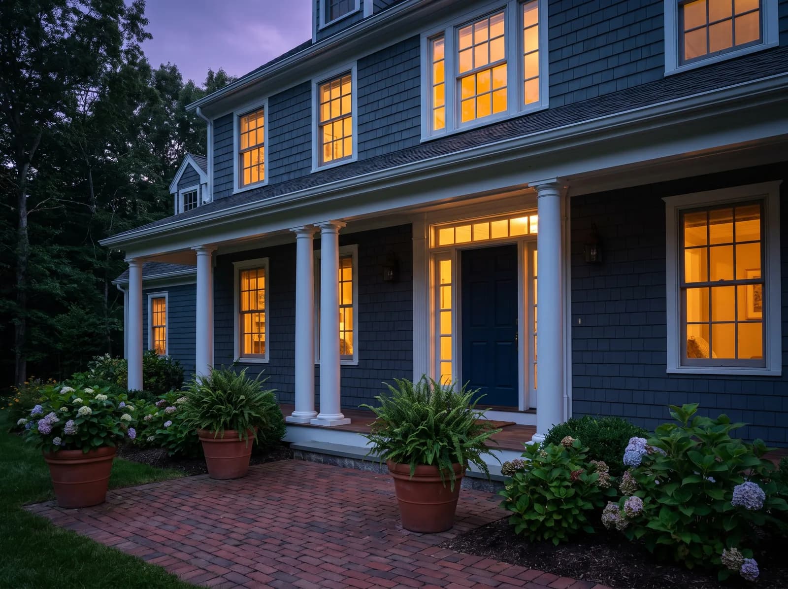 The covered front entrance of a shingled house at dusk, with the interior lights on